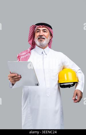Saudi senior man wearing traditional dishdasha and ghutra, holding a digital tablet and carrying a yellow hard hat, representing construction, technol Stock Photo