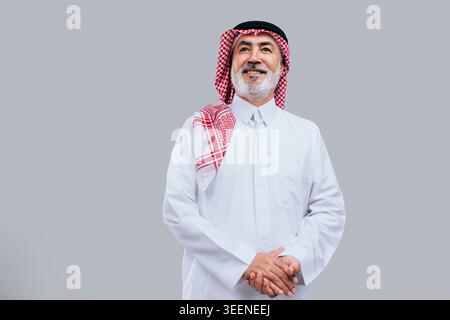 Saudi senior man wearing traditional ghutra and thobe, standing with hands clasped, smiling gently, and looking up, radiating a confident and optimist Stock Photo