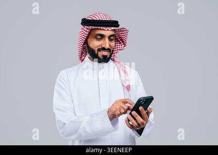 Saudi man in traditional thobe and ghutra smiling while using his smartphone, illustrating modern digital communication, connectivity and mobile techn Stock Photo
