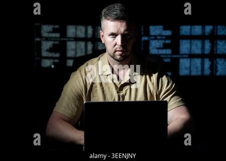 Hacker works late in dark room. Hacker analyzes software and programming code. Hacker monitors secure server system. Hacker creates ransomware attack Stock Photo