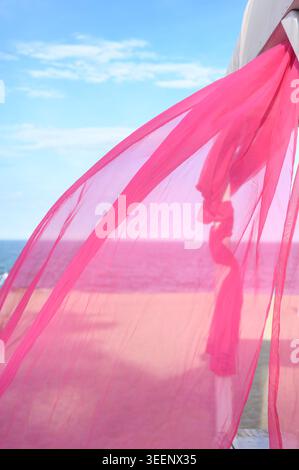 Light pink textile blowing in the wind against a maritime horizon. Soft ripples reach the sand. Concept of aesthetic vacation and natural beauty Stock Photo