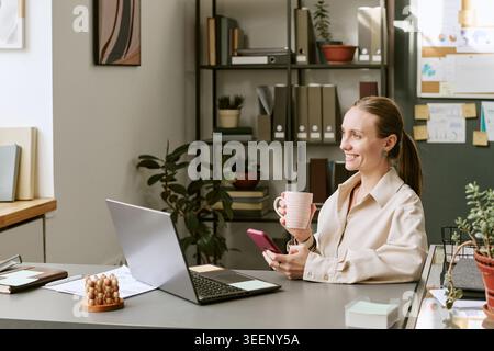 Adult Woman Using Smartphone and Drinking Coffee at Work Stock Photo