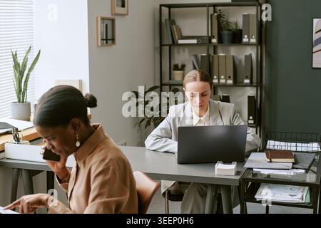 Black Woman Calling Client While Woman Working on Laptop in Office Stock Photo