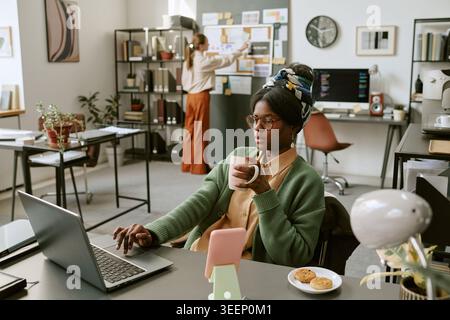 Black Woman Working on Laptop While Drinking Coffee in Office Stock Photo