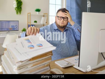 Stressed accountant at desk with paperwork stack and charts on computer screen Stock Photo