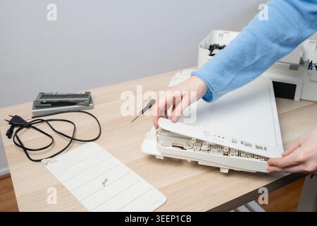Hands of a technician disassembling a flatbed scanner printer on a wooden desk with tools, cables and spare parts, illustrating office equipment repai Stock Photo