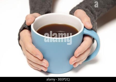 Close-up of human hands in a grey knitted sweater holding a large blue ceramic mug filled with black coffee isolated on a white background Stock Photo