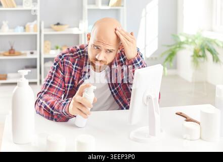 Bald man looking in the mirror sitting at table with cosmetics bottles at home. Stock Photo