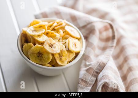 A white bowl filled with crunchy banana chips sits on a bright wooden table beside a soft checked kitchen towel, evoking homemade cooking, healthy sna Stock Photo