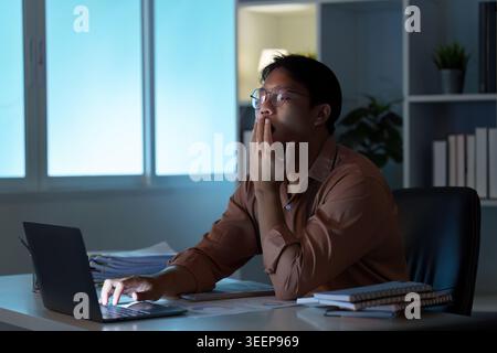 Tired Asian man yawning while working on laptop at remote home office at night Stock Photo