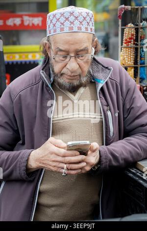An Americanized Muslim man wearing a Kufi prayer hat & remnants of a henna beard and also using airpods at a stand on 37th Ave in Jackson Heights, NYC. Stock Photo