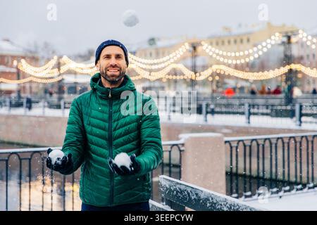 Pleasant looking male model keeps snowballs in hands going to have battle, waits for friends, looks happily into distance, being in good mood, stands outdoors. Beautiful winter views Stock Photo
