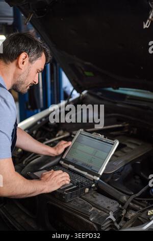 Car mechanic using diagnostic software on laptop, examining engine in auto repair shop Stock Photo