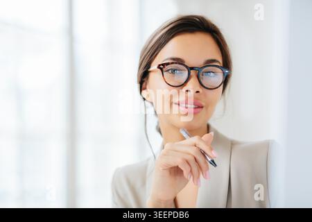 Headshor of attractive brunette woman holds pen, looks confidently at camera through spectacles, dressed in formal clothes, writes down notes, poses indoor. People, business and work concept Stock Photo