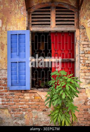 A red and blue shuttered rustic window tucked away in the back streets of Ipoh, framed by a weathered brick wall. Stock Photo
