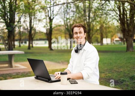 A young man smiles while working on a laptop at a stone table in a park. He wears headphones around his neck, enjoying a remote work lifestyle outdoor Stock Photo