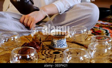 Detailed shot of a teapot pouring hot water into a ceramic cup on an ornate wooden tray surrounded by empty glass cups. Mindful tea rituals create Stock Photo