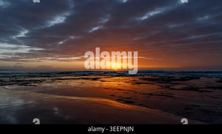 Aerial view of the sun setting over the rocky coastline of Hartenbos beach with dramatic clouds and golden reflections on the water Mossel Bay, Wester Stock Photo