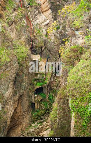 Old concrete platforms, staircases, and pipes cling to the steep rock walls of a narrow green gorge in Ronda, Andalusia, Spain. Moss and vegetation gr Stock Photo