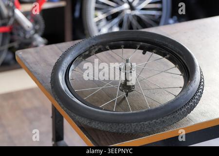Bicycle wheel with a deflated rubber tire resting on a wooden table, suggesting flat tire repair, maintenance, or servicing in a bike workshop, repres Stock Photo