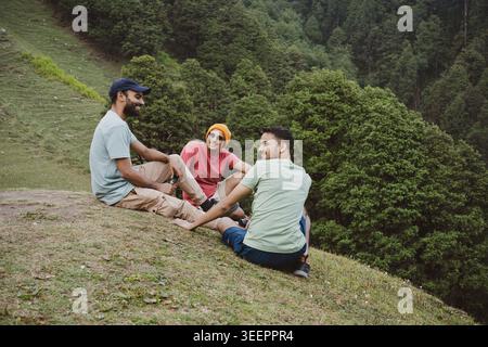 Three young friends sitting on a grassy slope overlooking a forest at Raghupur Fort, Sojha, Manali, Himachal Pradesh, India. Stock Photo