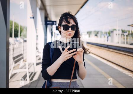 Woman stands on railway platform while using smartphone. Empowered traveler lifestyle, solo trip planning, confident city explorer Stock Photo