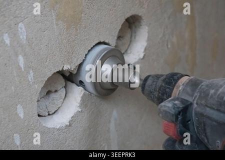 Worker drilling a large circular hole in a rough concrete wall using an electric hammer drill with a core bit, creating openings for electrical boxes Stock Photo