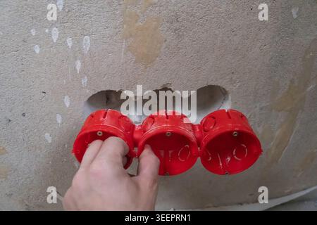 Worker's hand installing multiple red plastic electrical wiring boxes into a recessed hole in a rough concrete wall during a home renovation or constr Stock Photo