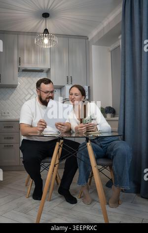 Young couple sitting at a glass table in a modern kitchen reviewing documents and calculating money, feeling stress and worry while discussing househo Stock Photo