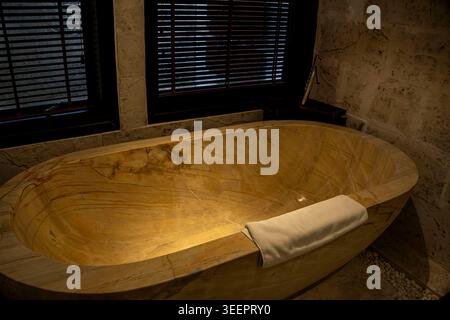 Stunning freestanding onyx stone bathtub with golden veining and white towel in a luxury resort bathroom in Vietnam. Dark wood blinds, marble walls, a Stock Photo