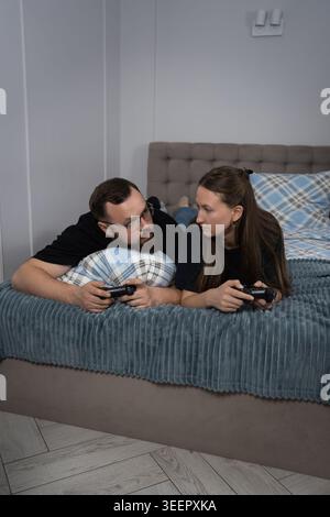 Young couple lying in bed holding game controllers in their hands and focused on competitive console game looking at each other while enjoying relaxed Stock Photo