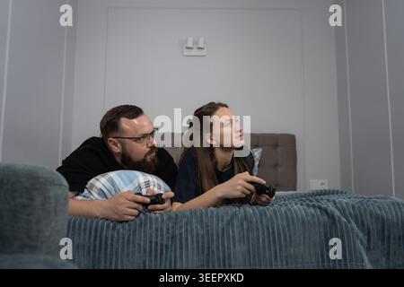Young heterosexual couple lying comfortably in bed, playing video games with wireless controllers, experiencing leisure, entertainment, and relaxation Stock Photo