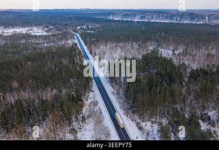 Aerial view of a straight asphalt road cutting through a dense snow-covered pine forest with a truck and cars in winter Lithuania. Stock Photo