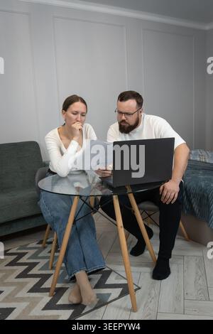 Young couple discussing financial documents, managing personal budget, and making online payments using a laptop in their home living space, focusing Stock Photo