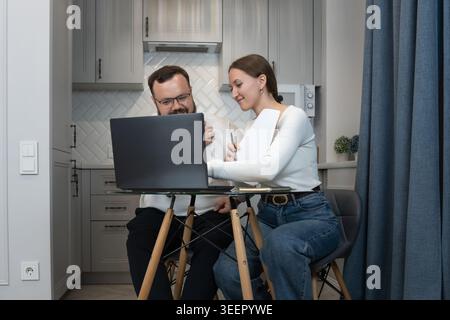 Young happy couple collaborating at a glass kitchen table, smiling while using a laptop and notebook to plan projects, manage finances and study toget Stock Photo