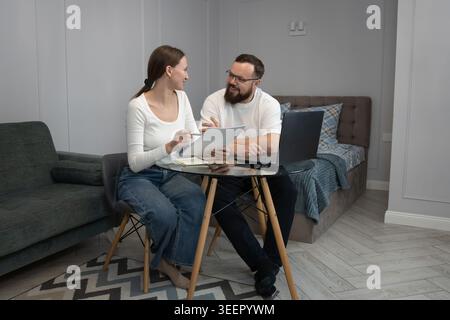 Young adult couple collaborating and sharing ideas, analyzing financial papers and using a laptop while sitting at a glass table in their modern home Stock Photo
