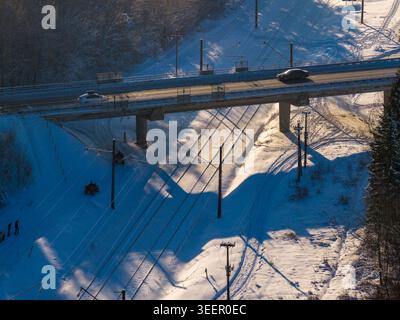 Aerial view of a road bridge crossing over snow-covered railway tracks with people and quad bikes in a wintry forest with long shadows in Lithuania. Stock Photo
