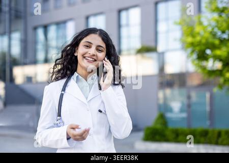 Young female doctor with a stethoscope wearing a white lab coat talking on a mobile phone and smiling, standing outdoors in front of a modern building Stock Photo