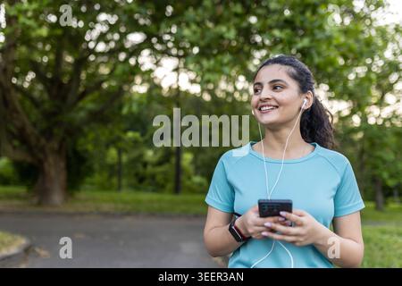 Young woman listening to music through wired earphones on her smartphone, wearing a fitness tracker and a sports t-shirt, smiling happily in a blurred green park environment Stock Photo