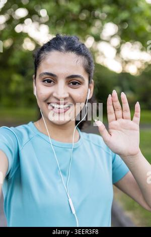 Young woman smiling at the camera and waving her hand, wearing a blue sports shirt and earphones while enjoying her outdoor fitness routine in a park setting Stock Photo