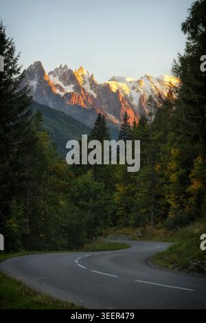 Aुberge La Boerne, France - 18.09.2025: A curving asphalt road leads through a coniferous forest toward sunlit, jagged alpine peaks at golden hour. Stock Photo