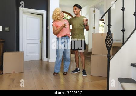 Diverse couple standing in foyer holding takeaway coffee cups beside stacked cardboard moving boxes Stock Photo