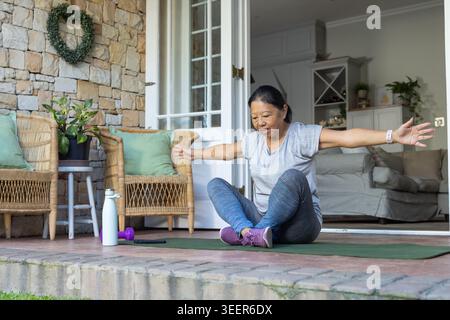 Senior Asian woman sitting on porch mat raising arms in gray tee near white bottle purple-dumbbell Stock Photo