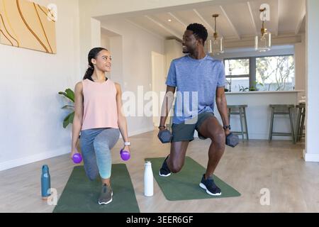 African American couple in sportswear doing forward lunges on green mats at home holding dumbbells Stock Photo