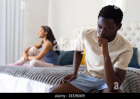 Couple sitting on bed in bedroom, man leaning forward holding chin, woman hugging knees, copy space Stock Photo