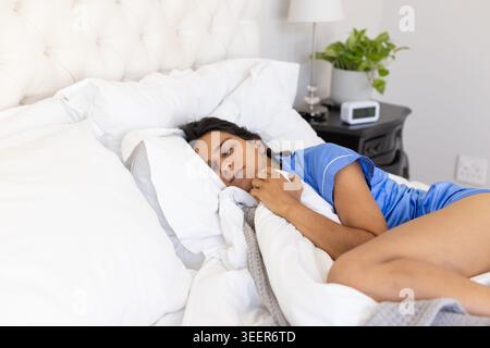 Indian woman sleeping on side in bedroom wearing blue pajamas on tufted bed beside nightstand lamp Stock Photo
