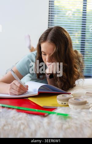 Female lying on stomach writing in open notebook on bed with folders, ruler, headphones Stock Photo