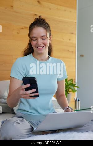 Woman sitting on bed, holding smartphone while using laptop, wearing light blue T shirt Stock Photo