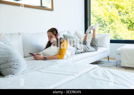 Female lying on white sofa, using phone, wearing pink headphones, smartwatch in living room Stock Photo