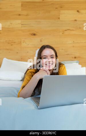 Female lying on stomach on bed wearing mustard yellow top, smiling and typing on silver laptop Stock Photo